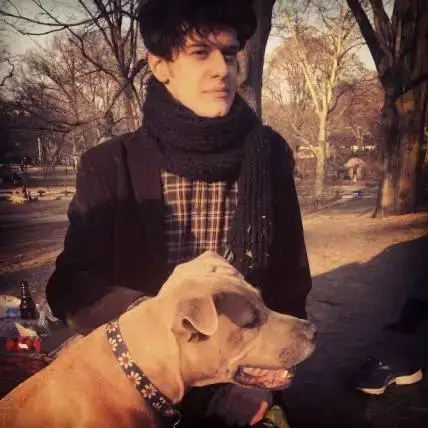 James Ortiz sits in a park wearing a thick black scarf and a plaid shirt while petting a tan dog with a floral collar.
