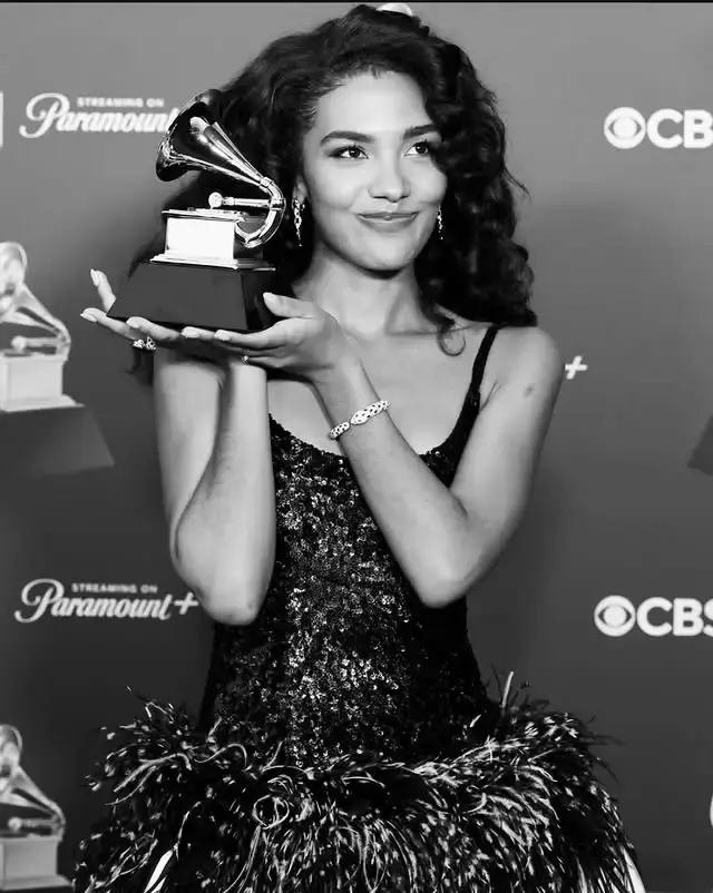 Olivia Dean smiles while holding a Grammy Award in a black-and-white photo, wearing a sequined dress with a feathered skirt.