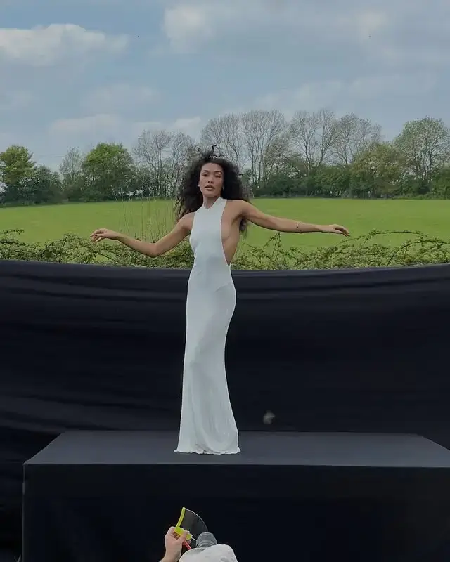 Olivia Dean poses in a lush green field wearing a backless white dress that flows behind her as she looks over her shoulder.