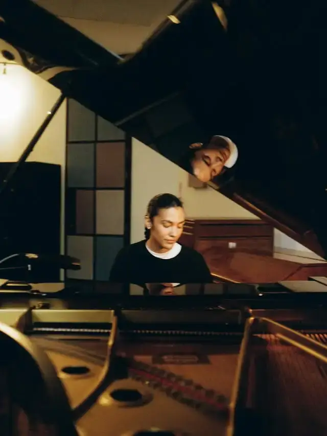 Olivia Dean sits focused at a grand piano with her reflection visible in the polished black lid of the instrument.