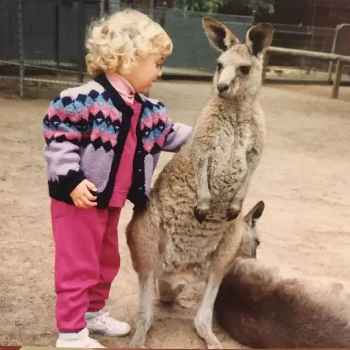 A young Liz Kingsman with blonde curly hair stands outdoors in a purple patterned cardigan and pink trousers next to a kangaroo.