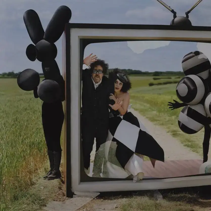 Tim Burton and Helena Bonham Carter pose whimsically inside a large, oversized television frame set in an open field for a Vogue photoshoot.