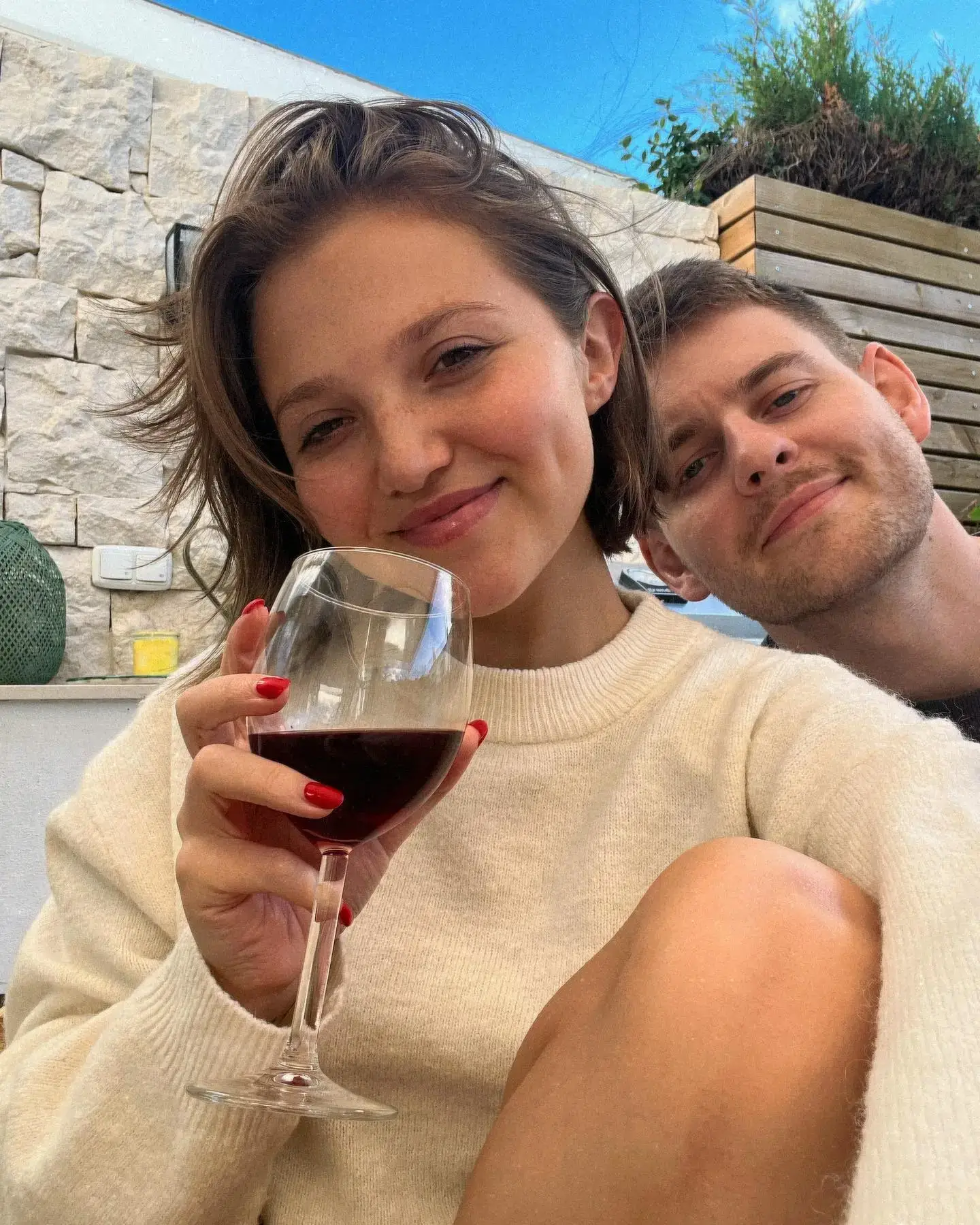 Mia McKenna-Bruce and Tom Leach smile for a close-up selfie while relaxing outdoors, with Mia holding a glass of red wine.