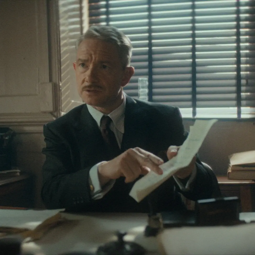Martin Freeman leans over a desk while holding a document and looking intently at something off-camera in a scene from Agatha Christie’s Seven Dials.