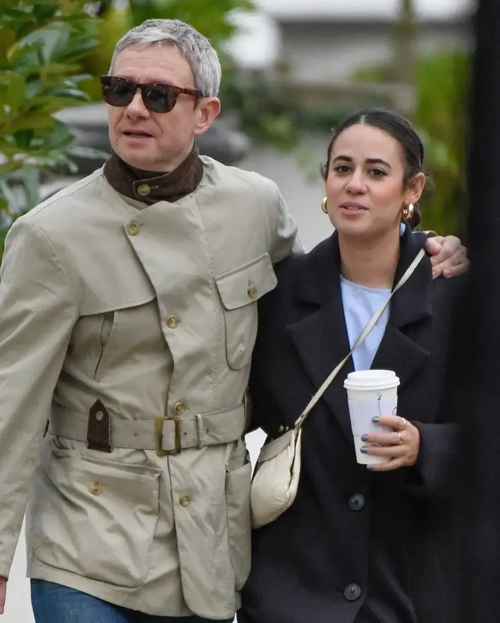 Martin Freeman and Rachel Mariam walk together outdoors, with Martin in a belted tan safari jacket and Rachel in a black coat holding a coffee cup.