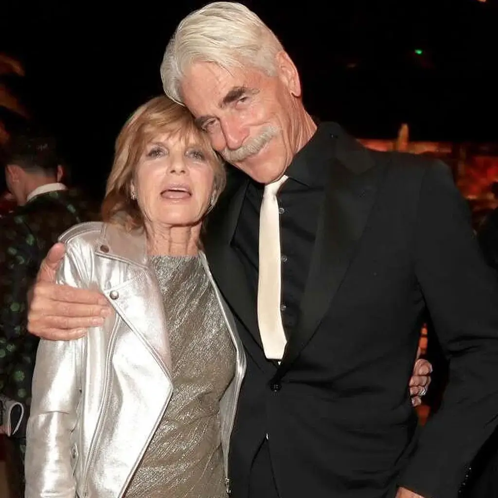 Sam Elliott and Katharine Ross pose closely together at an event, with Sam in a black suit and white tie and Katharine wearing a shimmering dress and silver leather jacket.