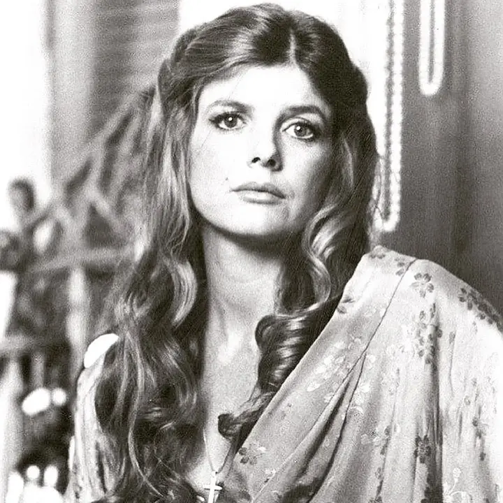 A vintage black and white portrait of Katharine Ross with long, wavy hair wearing a floral patterned robe and a cross necklace.