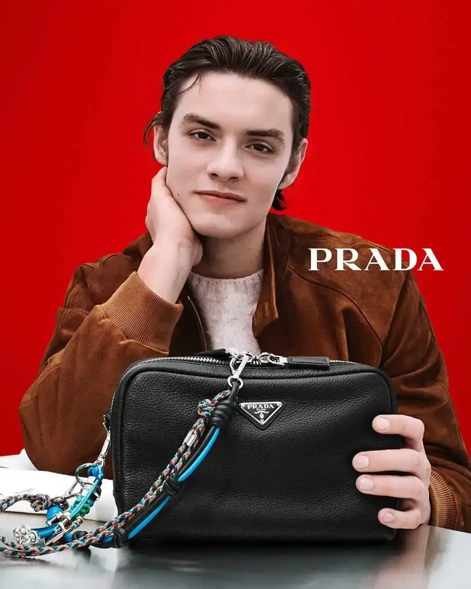 Louis Partridge leans his head on his hand while posing with a black leather Prada handbag against a vibrant red background.