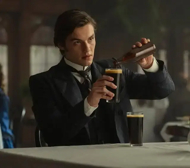 Louis Partridge stands in a 19th-century setting as Edward Guinness, wearing a formal Victorian suit with a high collar and a dark waistcoat.