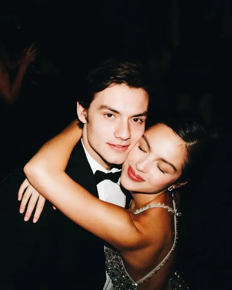 Louis Partridge and Olivia Rodrigo smile with Olivia in a red and white gingham dress and Louis in a blue shirt and burgundy tie.