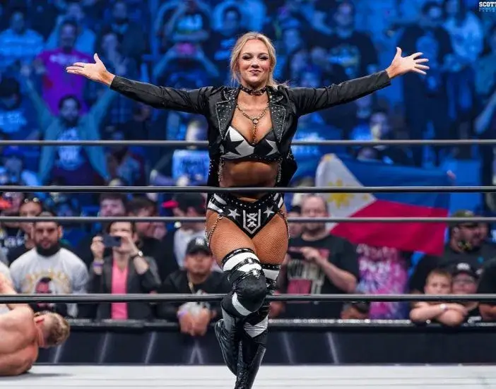 Anna Jay stands confidently in a wrestling ring with her arms outstretched, wearing star-patterned black and white gear and a studded jacket.