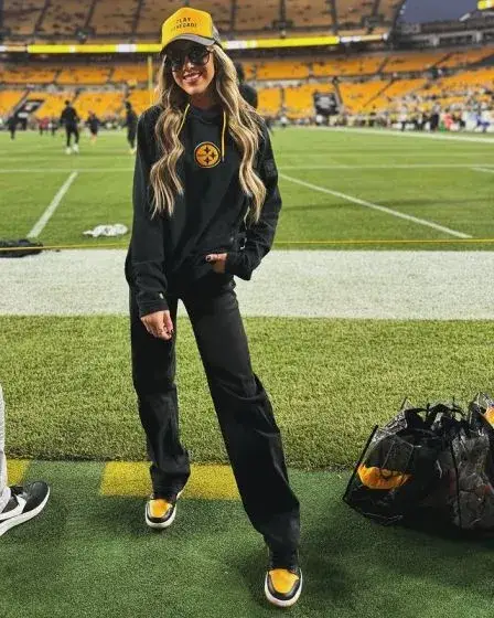 Professional wrestler Britt Baker stands on an American football field wearing a black Pittsburgh Steelers hoodie, matching pants, and a yellow hat.