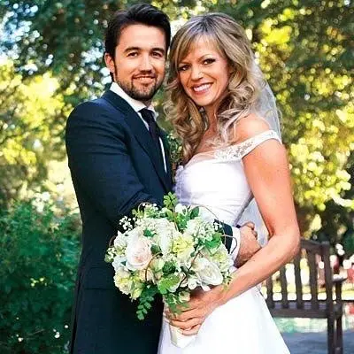 Rob McElhenney and Kaitlin Olson smile together in their wedding attire, with Olson holding a white and green floral bouquet against a lush green outdoor background.