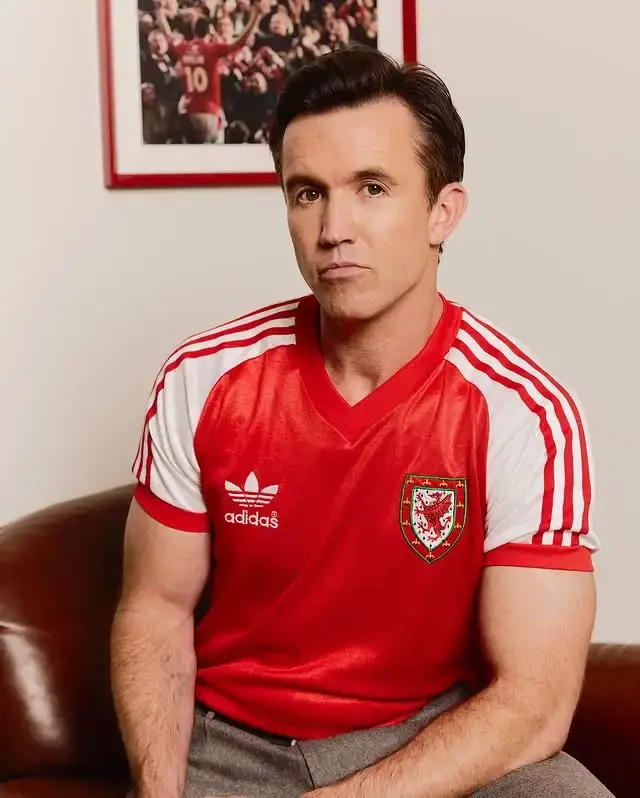 Actor and Wrexham AFC co-owner Rob McElhenney sits on a leather sofa wearing a red and white vintage-style Adidas football jersey featuring the Welsh dragon crest.