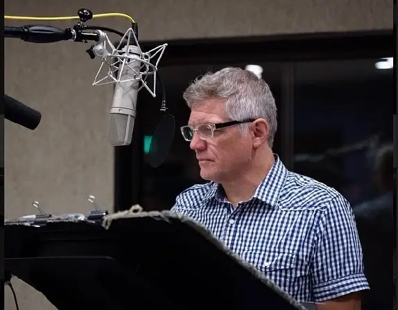 Actor Matthew Glave stands in a recording booth wearing a blue checkered shirt and glasses while looking down at a script near a professional microphone.