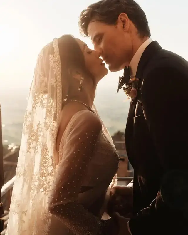 Riya Ray in a lace wedding veil and Enver Gjokaj in a tuxedo share an intimate moment outdoors during their wedding ceremony.