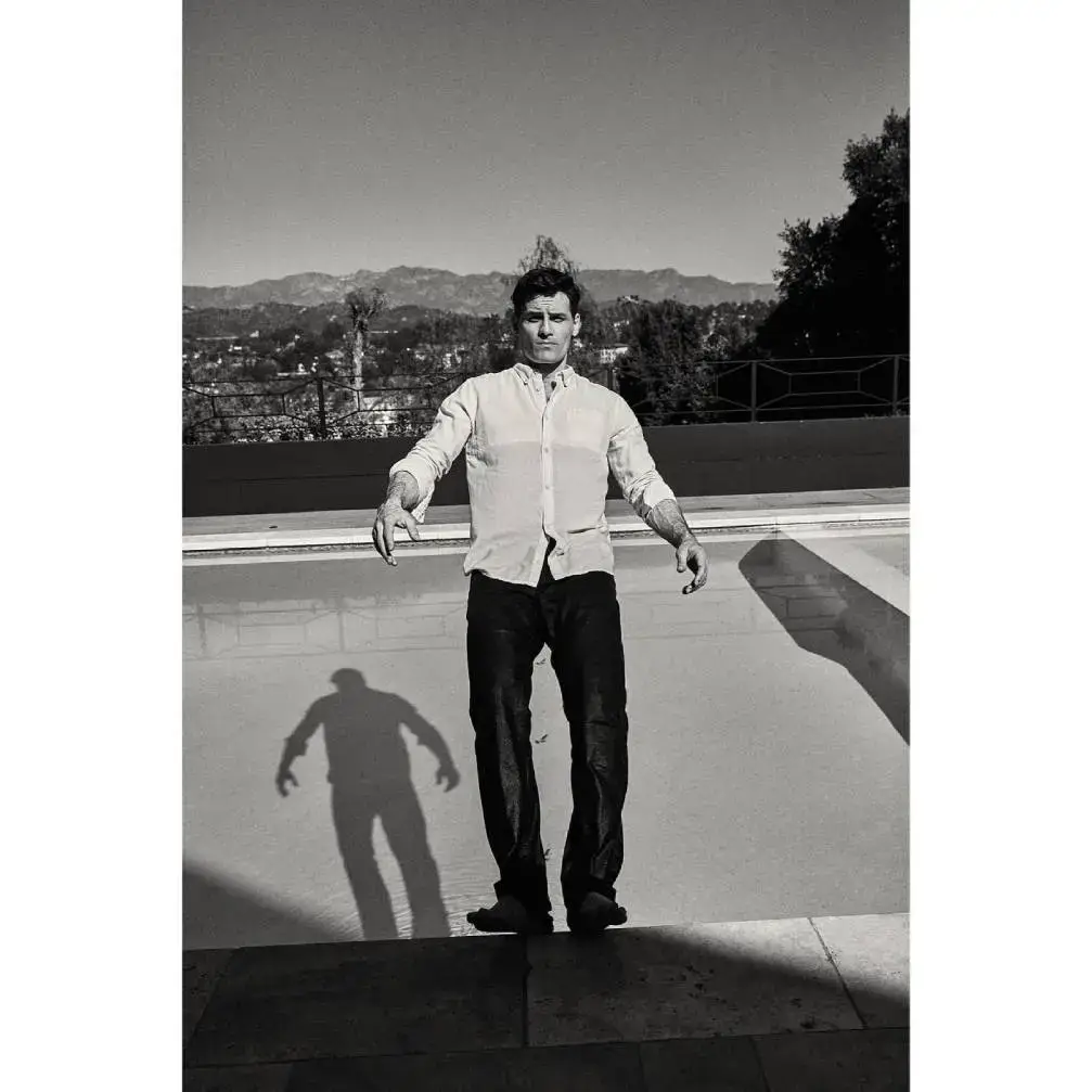 A black and white photo of actor Enver Gjokaj standing at the edge of a swimming pool with a scenic mountain range in the background.