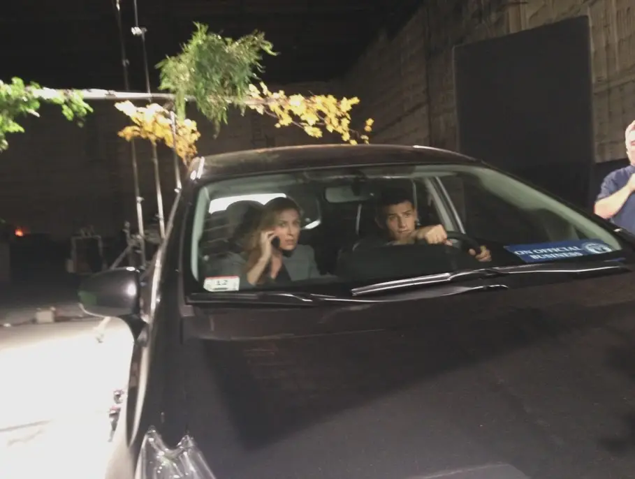 Actors Sasha Alexander and Enver Gjokaj sit inside a dark vehicle on a television set with production equipment and artificial foliage visible in the background.