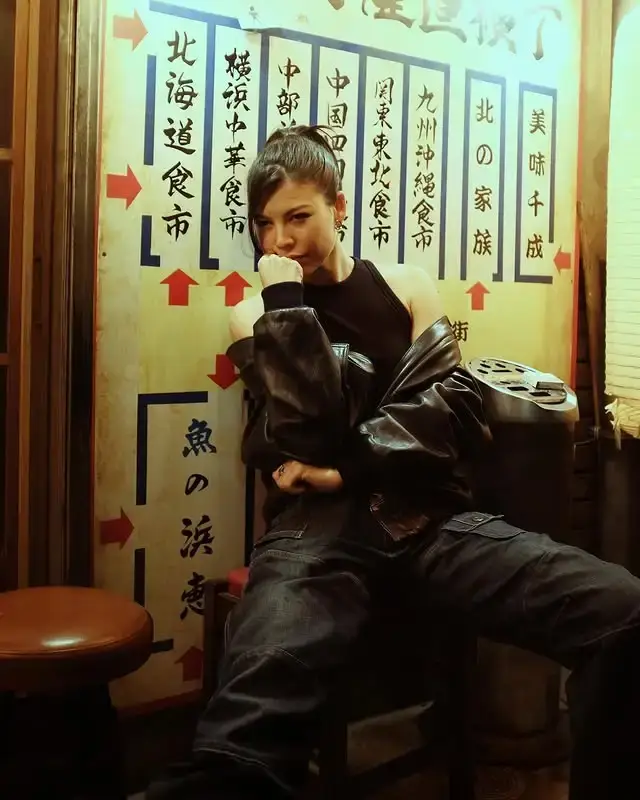 Sea Shimooka, an actress of Japanese, Native Hawaiian, French, and English descent, poses in a black leather jacket and cargo pants against a backdrop featuring Japanese text.