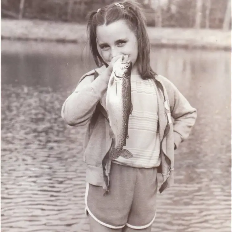A young Julie Ann Emery stands by a lake, proudly holding up a large fish she caught while wearing pigtails and a zip-up hoodie.