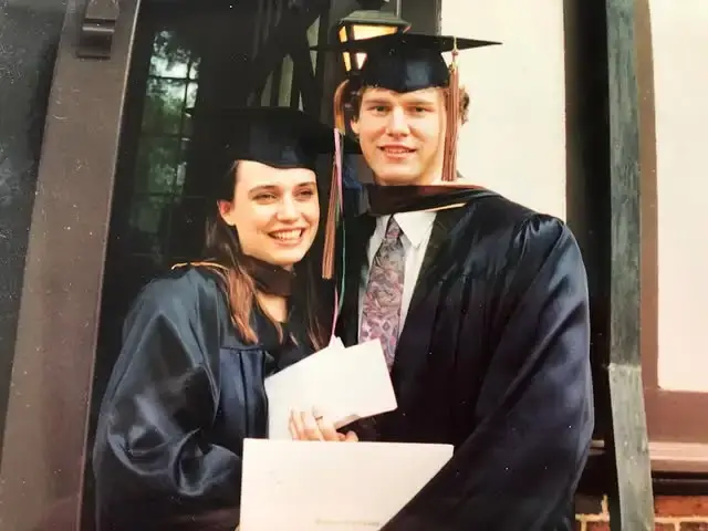 Kevin Earley and Julie Ann Emery smile together in their graduation caps and gowns while holding their diplomas from Webster Conservatory.