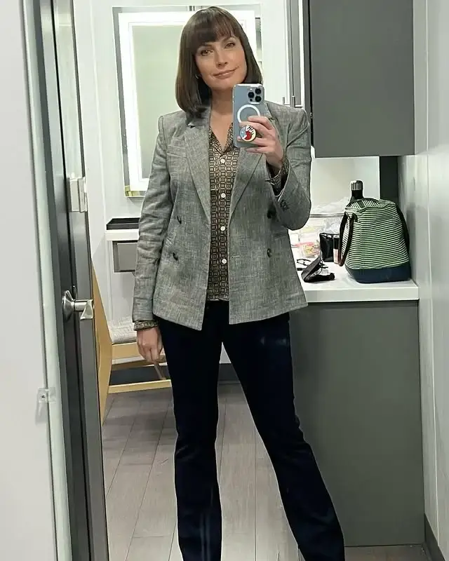 Actress Julie Ann Emery takes a full-length mirror selfie in a dressing room while wearing a grey blazer, patterned shirt, and dark trousers.