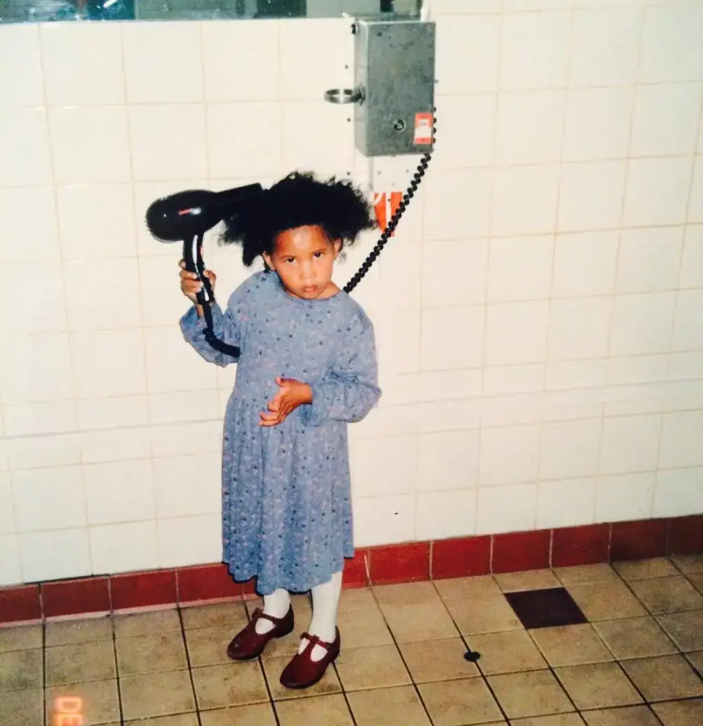 A childhood photo of Fola Evans-Akingbola as a young girl in a blue floral dress holding a hairdryer in a tiled room.