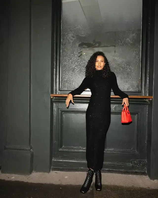 Actress Fola Evans-Akingbola posing in a long black turtleneck dress and lace-up boots while leaning against a dark wall and holding a small red handbag.
