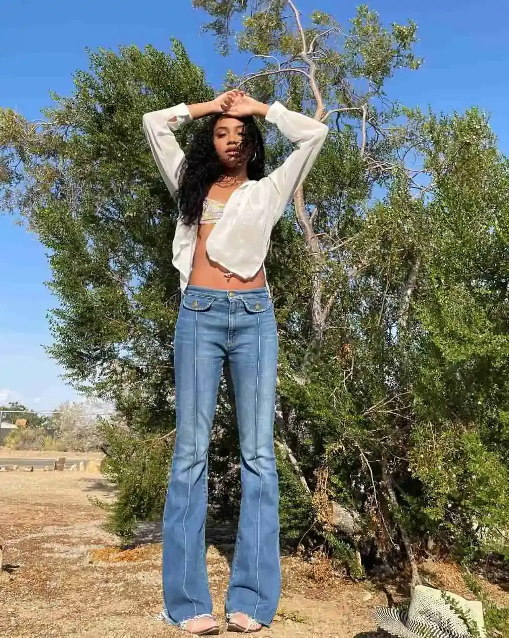 Genneya Walton poses outdoors wearing a white sheer button-down shirt over a bikini top and blue flared jeans with her arms raised above her head.