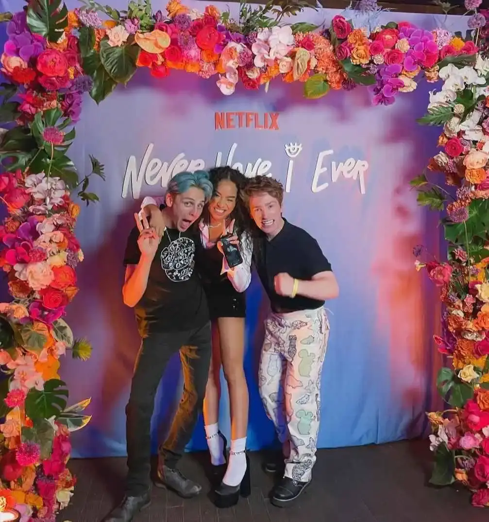 Jacob Bertrand, Genneya Walton, and Cooper Roth smile and pose together in front of a blue "Never Have I Ever" Netflix backdrop framed by a vibrant floral arch.