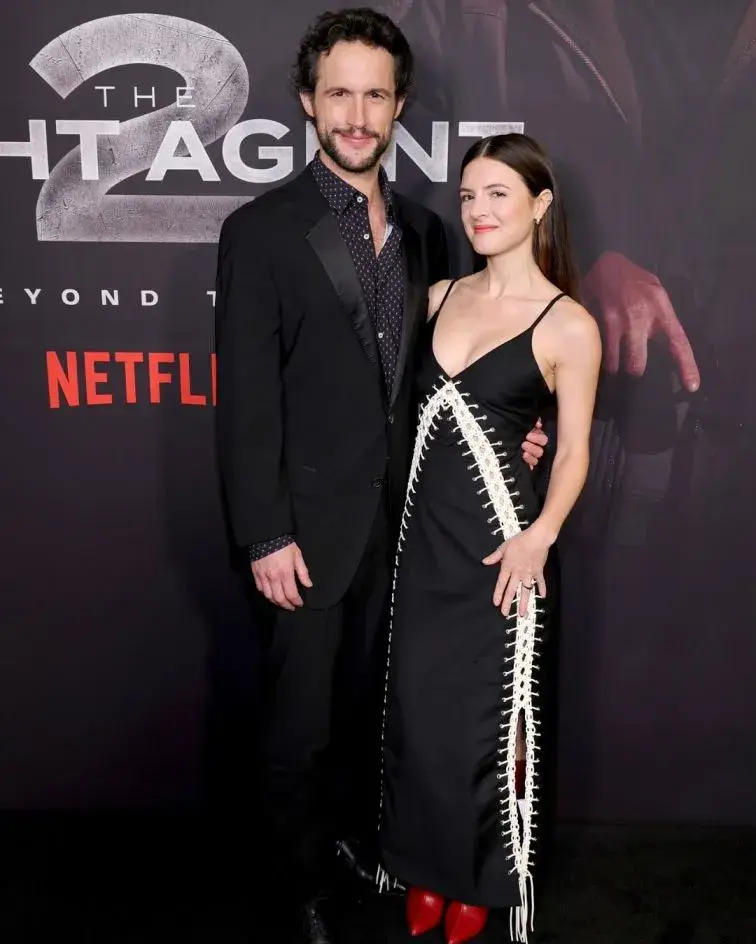 Rob Heaps in a black suit and Elise Kibler in a black dress with white lace-up detailing pose together on a red carpet in front of a Netflix "The Night Agent" backdrop.
