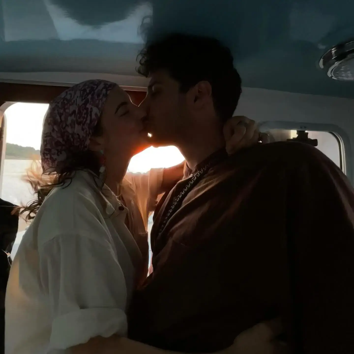 Elise Kibler, wearing a patterned headscarf and white shirt, shares a romantic kiss with her boyfriend inside a boat against the glow of a setting sun.
