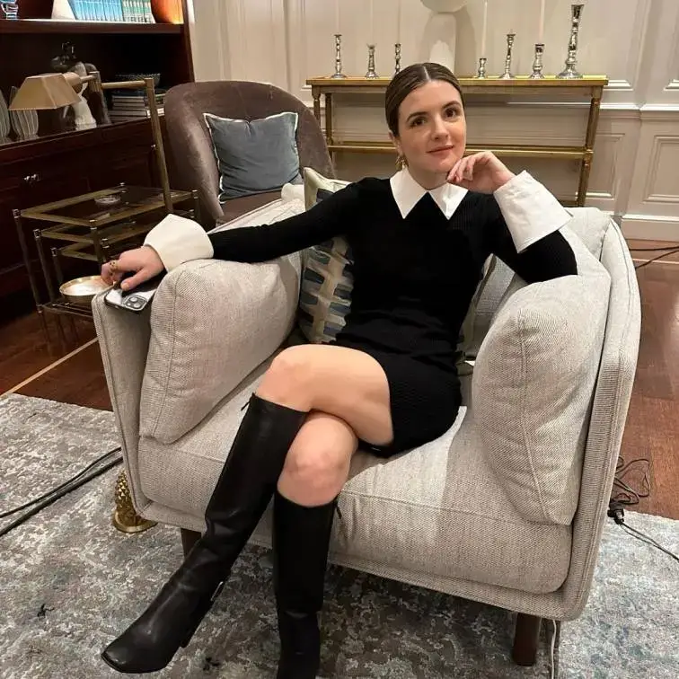 Elise Kibler sits with her legs crossed in a light gray armchair, wearing a long-sleeved black dress with a sharp white collar and cuffs paired with tall black boots.