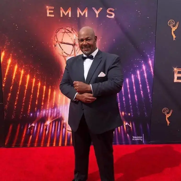 Kevin Michael Richardson poses confidently on a red carpet in a dark suit and bow tie at the Emmy Awards.