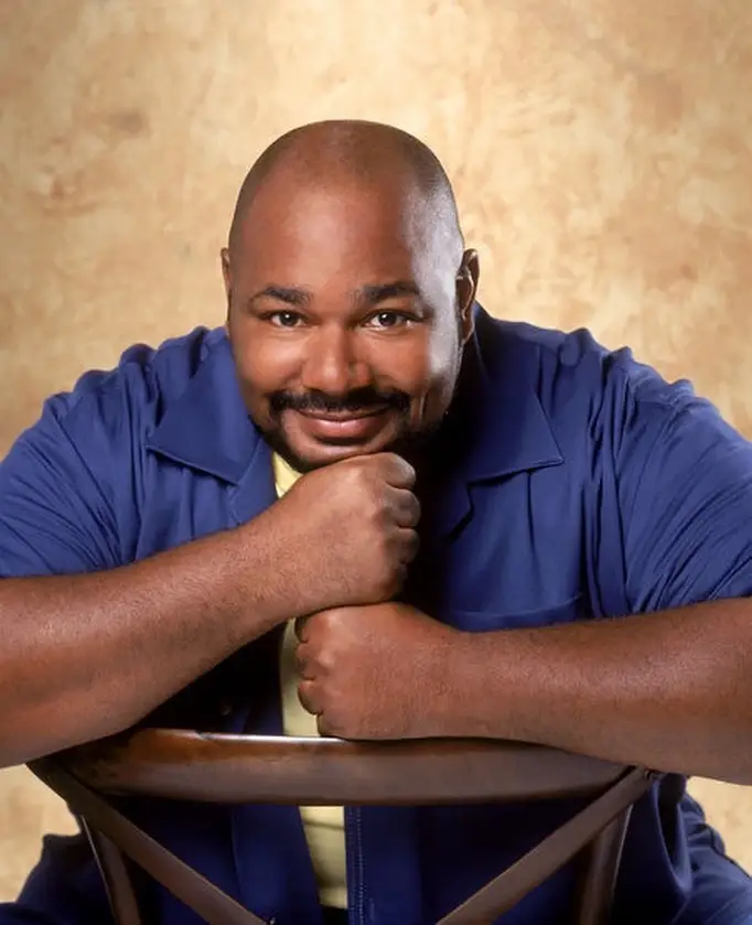 Voice actor Kevin Michael Richardson is captured in a headshot wearing a blue button-down shirt and smiling against a neutral backdrop.