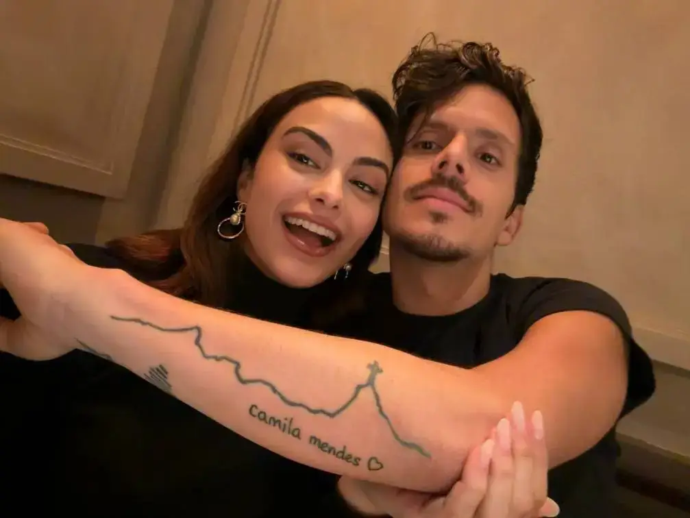 Camila Mendes and Rudy Mancuso pose together while Rudy displays a forearm tattoo that features the name "Camila Mendes" next to a heart and a mountain skyline.