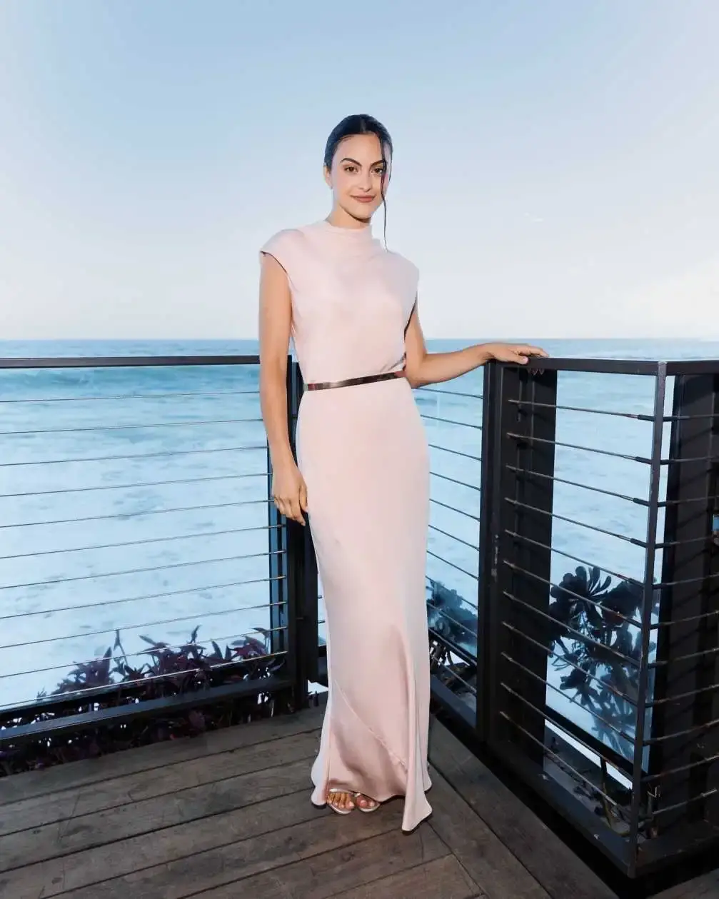 Camila Mendes poses on a wooden balcony overlooking the ocean, wearing a sleeveless, floor-length pale pink gown accented with a thin gold belt.