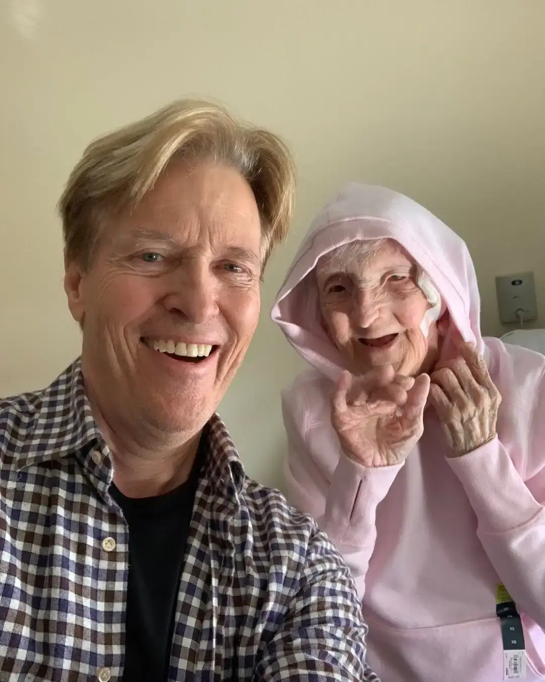 Actor Jack Wagner smiles warmly in a selfie next to his elderly mother, who is wearing a light pink hooded sweatshirt.