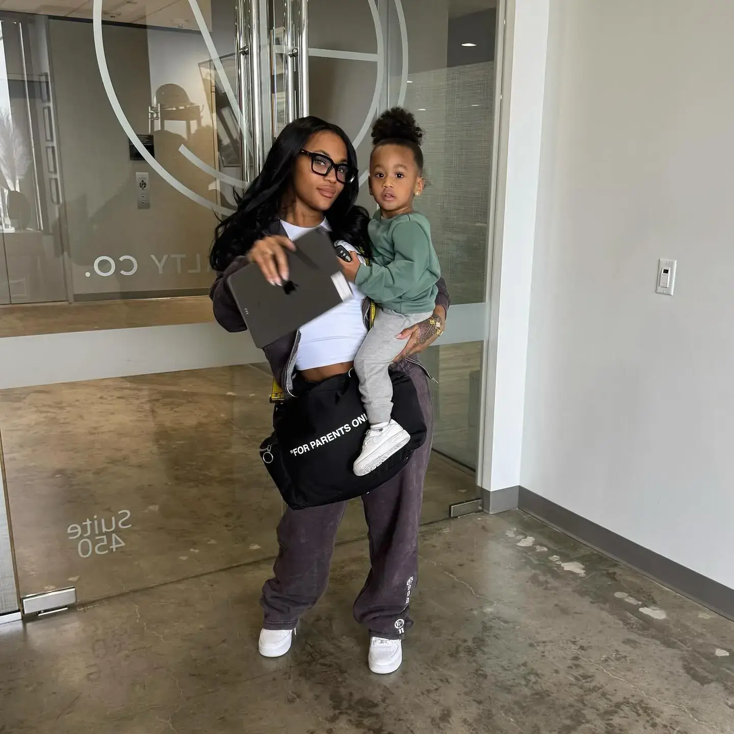 Lira Galore wears glasses and a brown tracksuit while holding her daughter Khaleesi, who is dressed in a green sweatshirt and grey joggers.