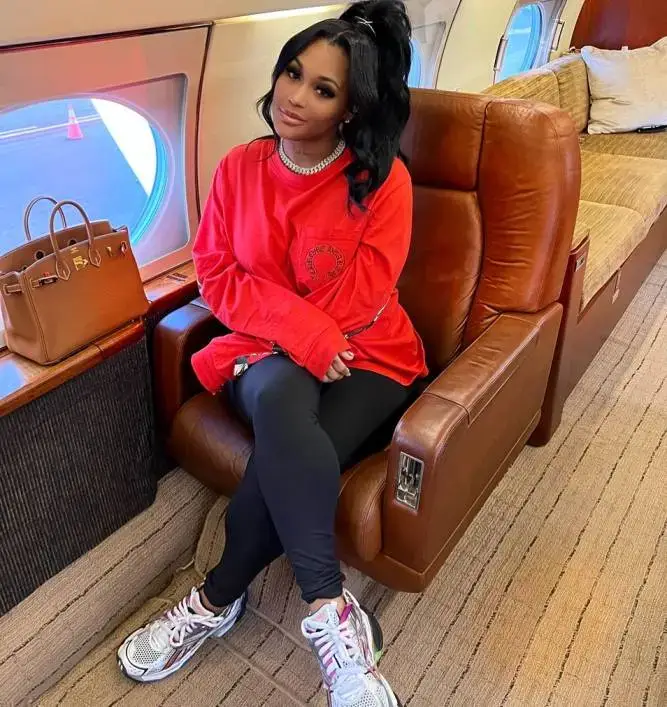 Lira Galore sits comfortably on a private jet wearing a bright red long-sleeve shirt, black leggings, and multi-colored sneakers.