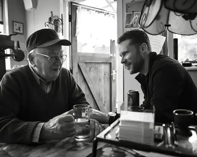 A black and white photo shows Jack Reynor smiling while sitting at a table across from Uncle Andy, who is wearing a flat cap and glasses.