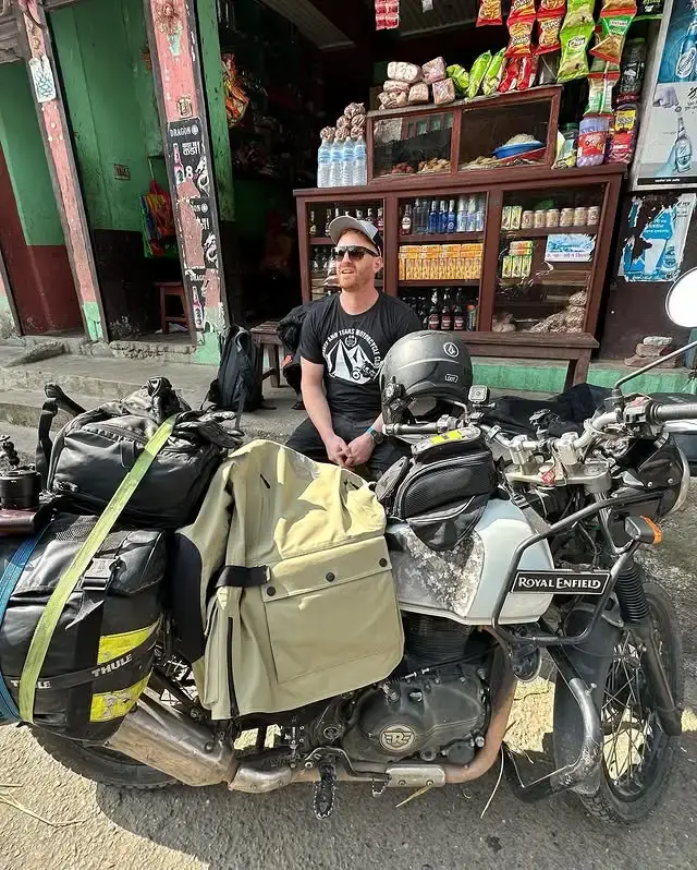 Actor Jack Reynor sits outside a roadside shop in Nepal next to a Royal Enfield motorcycle loaded with travel gear.