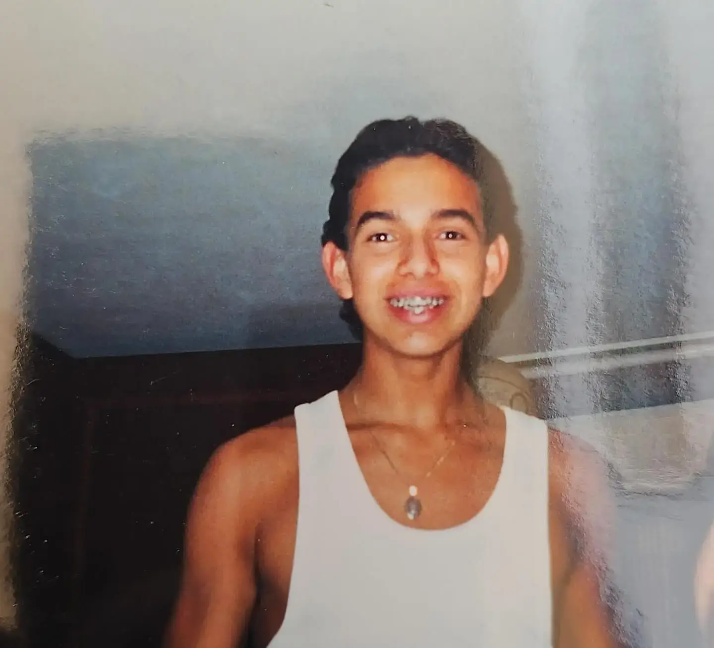 A young Alejandro Edda smiles for a childhood photo while wearing a white tank top and a necklace.