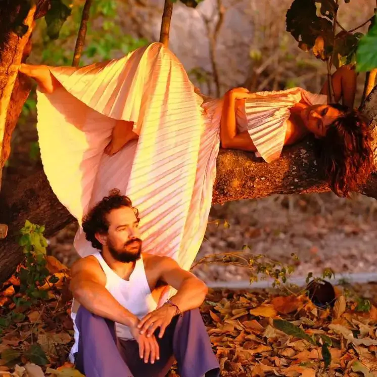 Niki Trosky lies gracefully on a large tree branch while Alejandro Edda sits on the leaf-covered ground beneath her during a golden hour photo shoot.