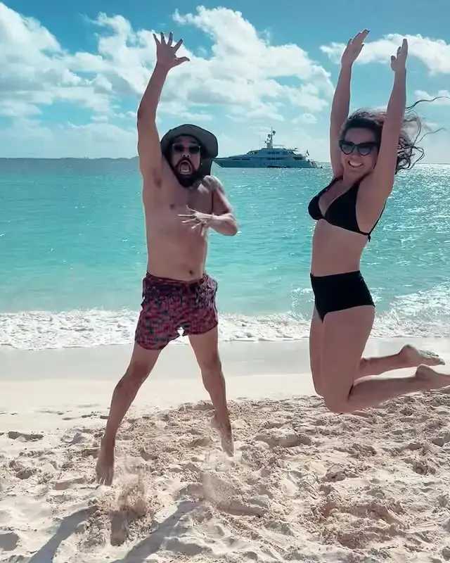 Alejandro Edda and his wife Aura Serna jump excitedly in the air on a white sand beach with turquoise water and a yacht in the background.