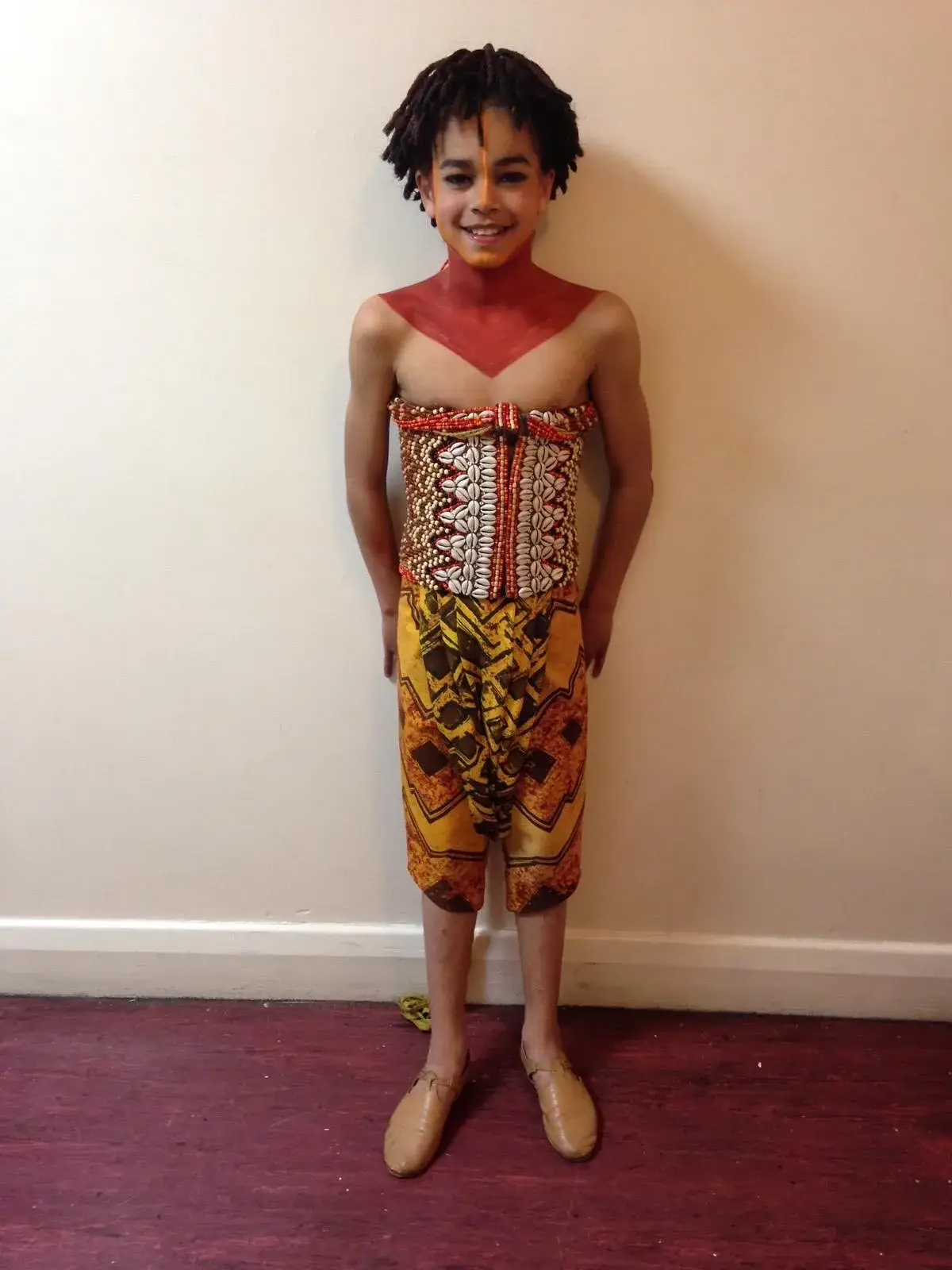 A young Amir Wilson poses in a vibrant stage costume featuring an ornate beaded corset, patterned yellow trousers, and red neck paint.