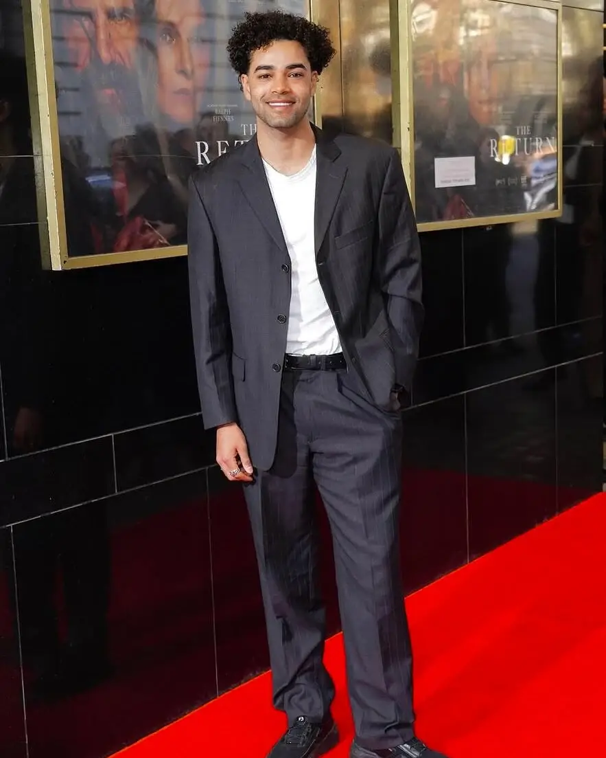 Actor Amir Wilson smiles on a red carpet while wearing a charcoal pinstripe suit over a white t-shirt.