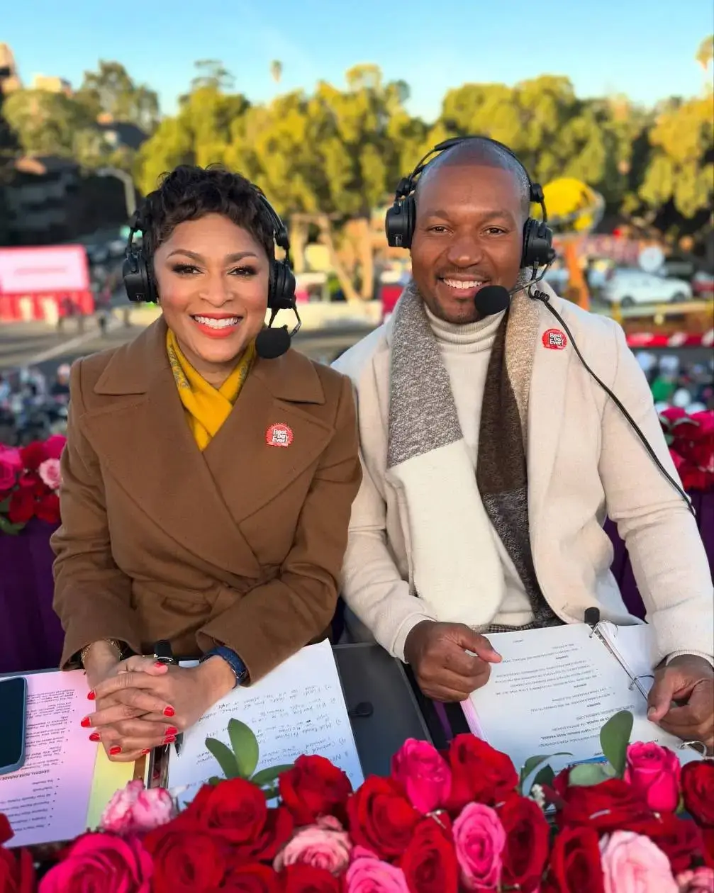 Broadcasters Jericka Duncan and Darren M. Haynes smile at a floral-decorated desk while wearing headsets during an outdoor event.