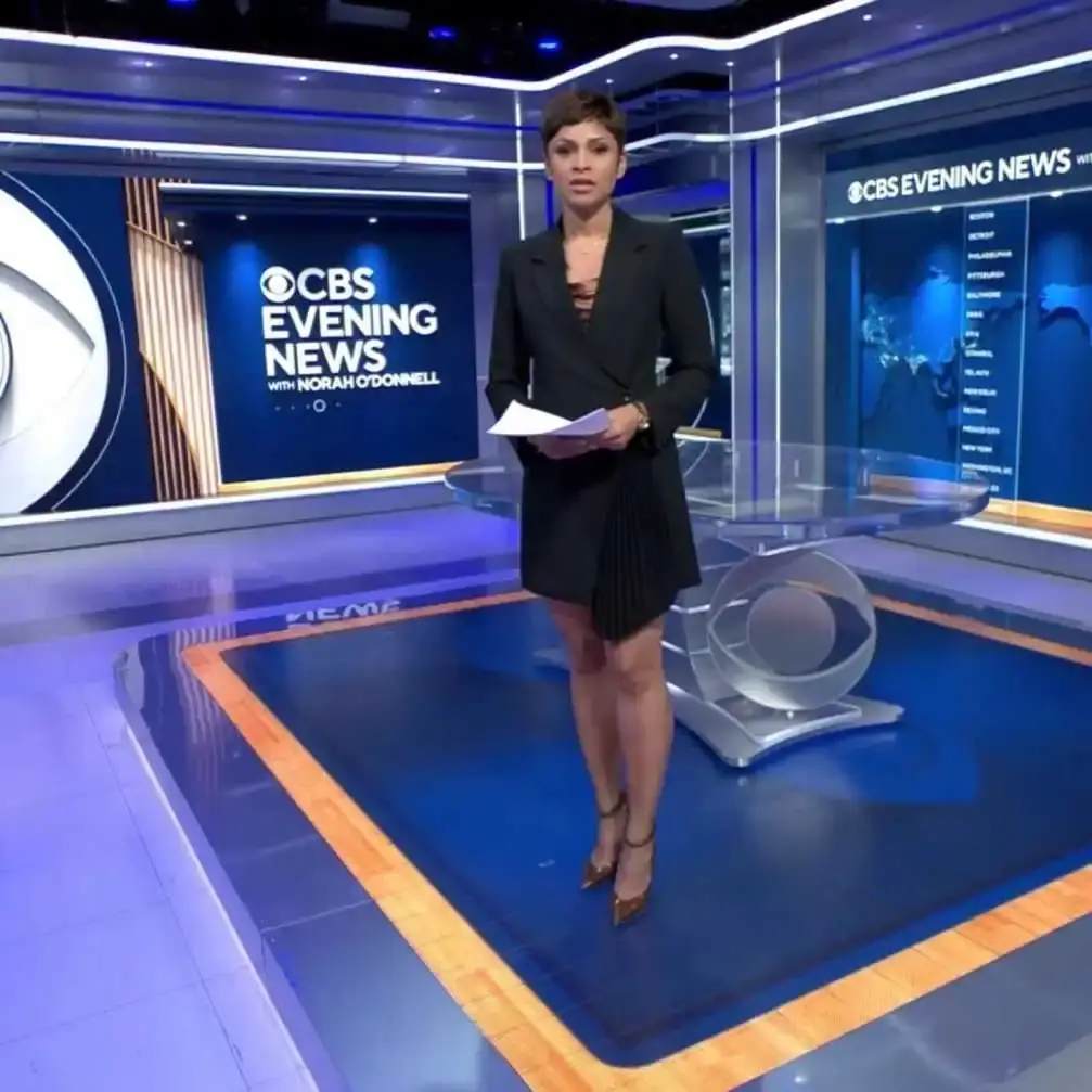 News anchor Jericka Duncan stands in a modern blue-lit studio wearing a black blazer dress while holding reporting scripts.