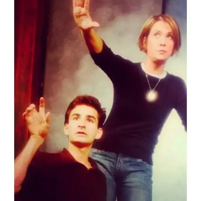A vintage photo of a young Kristen Wiig with short hair and a black sweater performing on stage alongside a male actor.